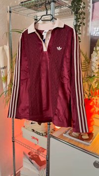 Μπορντώ αθλητική μπλούζα με γιακά, υφή σαν δαντέλα, και λευκό λογότυπο adidas. Διαθέτει τρεις λευκές ρίγες κατά μήκος κάθε μανικιού.