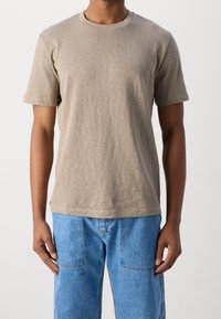 T-shirt beige in cotone con collo rotondo e maniche corte, caratterizzata da un sottile logo sul petto. Abbinata a jeans in denim blu.