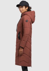 Brun, lang, polstret frakke med hætte, vertikale syninger og sidelommer. Har sorte knapper og snoretræk detaljer.