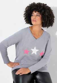 Grijze longsleeve t-shirt met een V-hals, featuring roze en witte sterontwerpen versierd met kleine strass-steentjes. Gecombineerd met zwarte leggings.