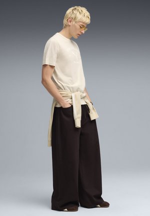 Beige katoenen T-shirt met logo, losse zwarte broek en licht beige sweater om de taille gestrikt; accessoires zijn onder andere gele zonnebril.