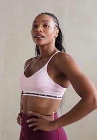 Sostén deportivo rosa con una banda contrastante en un oscuro color burdeos. Textura suave con tirantes finos. La modelo posa con leggings deportivos ajustados en color burdeos.