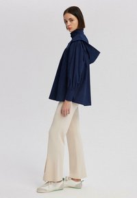 Tamno plava oversized majica s visokim okovratnikom i kapuljačom, uparena s kremasto bijelim hlačama koje se šire prema dnu i bijelim tenisicama s zelenim detaljima.