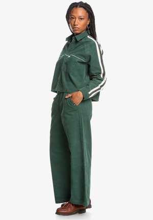 Green corduroy cropped jacket with front pocket and white stripe accents, paired with wide-leg pants in matching material. Brown lace-up shoes.