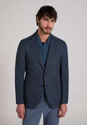 Man draagt een donkerblauw gestructureerd colbert over een blauwe poloshirt en lichtblauwe broek, staand tegen een effen lichte achtergrond.