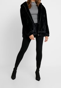 Schwarze Kunstfell-Jacke mit Reißverschluss und Kapuze, die eine weiche Textur und elastische Bündchen hat, kombiniert mit schwarzen engen Jeans und Stiefeletten mit Absatz.