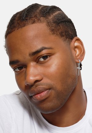Boucle d'oreille en argent avec un pendentif en croix, portée à l'oreille gauche d'une personne aux cheveux courts et tressés sur un fond neutre.