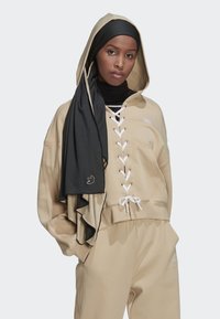 Hoodie beige corto con lacci sul davanti e legature bianche, abbinato a pantaloni coordinati. Un scarf nero testurizzato pende sopra il hoodie.