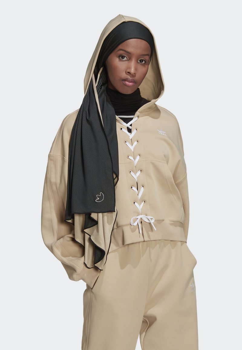 Hoodie beige corto con lacci sul davanti e legature bianche, abbinato a pantaloni coordinati. Un scarf nero testurizzato pende sopra il hoodie.