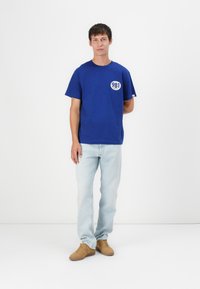 T-shirt in cotone blu a maniche corte, con un logo circolare bianco con il numero "981" sul lato sinistro del petto. Indossato con jeans chiari e scarpe marroni.