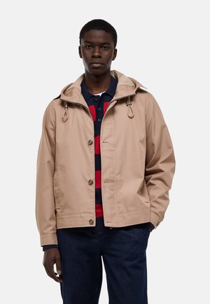 Jeune homme portant une veste à capuche beige par-dessus un polo rayé noir et rouge et un pantalon bleu foncé, debout devant un fond blanc.