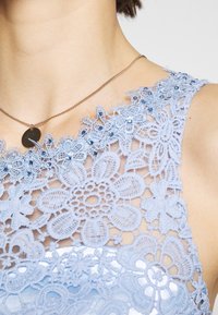 Gros plan d'une personne portant un haut en dentelle fleurie bleue orné de petites perles et un collier fin en chaîne dorée avec un pendentif rond.