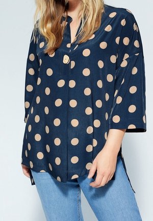 Mujer con blusa de manga larga azul marino con grandes lunares beige, combinada con vaqueros azul claro, posando con una mano cerca del muslo.