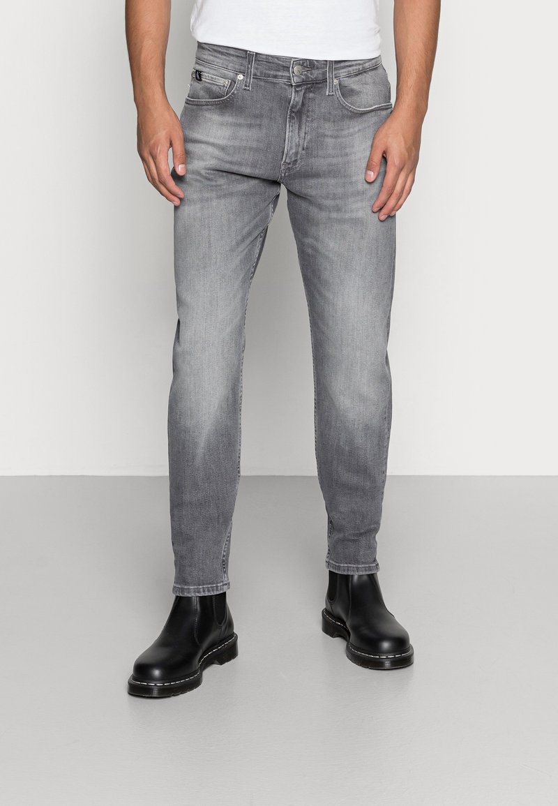 Jeans de mezclilla gris con corte recto, que presentan un diseño clásico de cinco bolsillos y un ligero desgaste, combinados con botas de cuero negro hasta el tobillo.