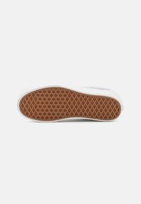 Semelle de chaussure avec motif gaufré en caoutchouc brun sur une semelle intermédiaire blanche sur fond blanc uni.