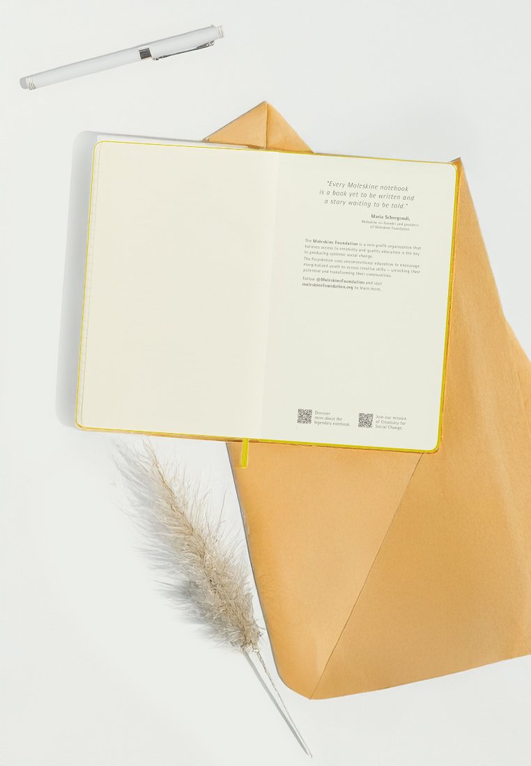 Moleskine-Notizbuch mit gelbem Rand, geöffnet auf leeren Seiten, neben einem braunen Umschlag und einer beigen Feder. Ein Stift liegt darüber.