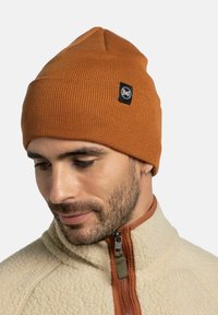 Gorro de punto en color óxido con textura de canalé y un parche de logo negro. Se lleva con una chaqueta de felpa color crema con cierre de cremallera.