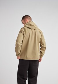 Chaqueta de lluvia beige con capucha, puños elásticos y un diseño elegante. La tela parece suave y resistente al agua. Pantalones negros al fondo.
