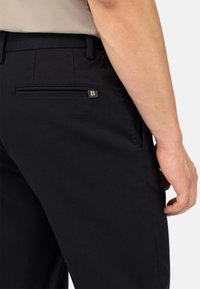 Juodos kelnės su užpakaliniu kišeniu su užtrauktuku ir metaliniu logotipo akcentu. Audinys turi tekstūruotą paviršių.