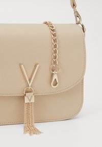 Beige syntetisk handväska med en gyllene kedjerem, en framträdande gyllene "V"-logotyp och en dekorativ tofs med kedjedetaljer.