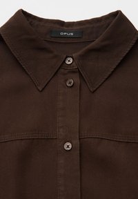 Gros plan sur une chemise boutonnée marron foncé avec un col pointu et une étiquette de marque « OPUS » visible à l’intérieur du col.