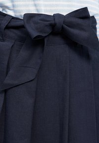 Mörkblå linnekjol med en rosettdetalj i midjan, med veck och en slät textur. Förstärkt med en stilren midjeband.