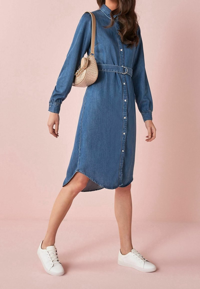 Robe-chemise en denim avec une taille ceinturée, des manches longues, une fermeture boutonnée à l'avant et un ourlet arrondi. Associée à des baskets blanches et un sac bandoulière couleur beige.