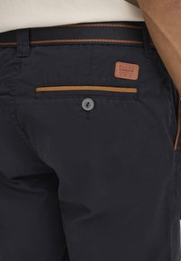 Pantalon bleu marine avec une texture lisse ; présente une étiquette en cuir marron, une fermeture à bouton, et une fine bande marron le long de la poche arrière.