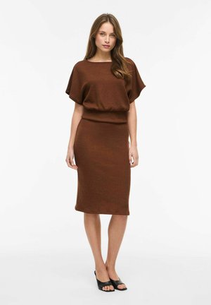 Žena v priliehavej hnedej šatke po kolená s krátkymi rukávmi a čiernymi otvorenými lodičkami stojaca na bielom pozadí.
