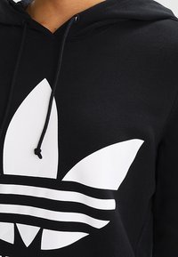 Svart hoodie med en framträdande vit logotyp med tre blad och tre horisontella ränder. Inkluderar dragkedjor vid halslinningen.