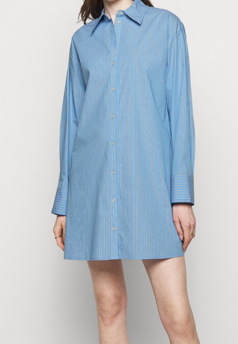 Femme portant une robe-chemise bleue à manches longues, boutonnée, avec des rayures blanches verticales et un col pointu.