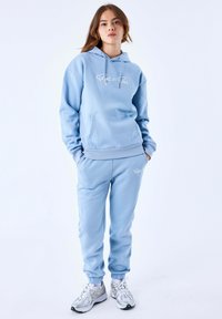 Felpa azzurra chiara con tasca frontale e logo bianco, abbinata a joggers coordinati con polsini elasticizzati e tasche laterali, indossati con sneakers.