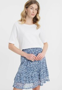 Witte top met korte mouwen gecombineerd met een blauwe rok met bloemenprint en ruches. De rok heeft een gelaagde structuur en een zachte, vloeiende stof.