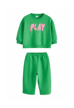 Zielona bluza z krótkim rękawem z różowym napisem "PLAY", zestawiona z zielonymi elastycznymi szortami na białym tle.