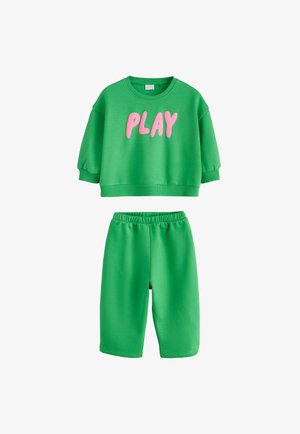Zielona bluza z krótkim rękawem z różowym napisem "PLAY", zestawiona z zielonymi elastycznymi szortami na białym tle.
