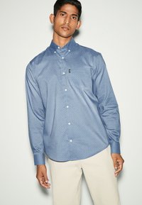 Camicia button-up a maniche lunghe con motivo blu, caratterizzata da un piccolo logo sul petto e un orlo arrotondato, abbinata a dei pantaloni chiari.