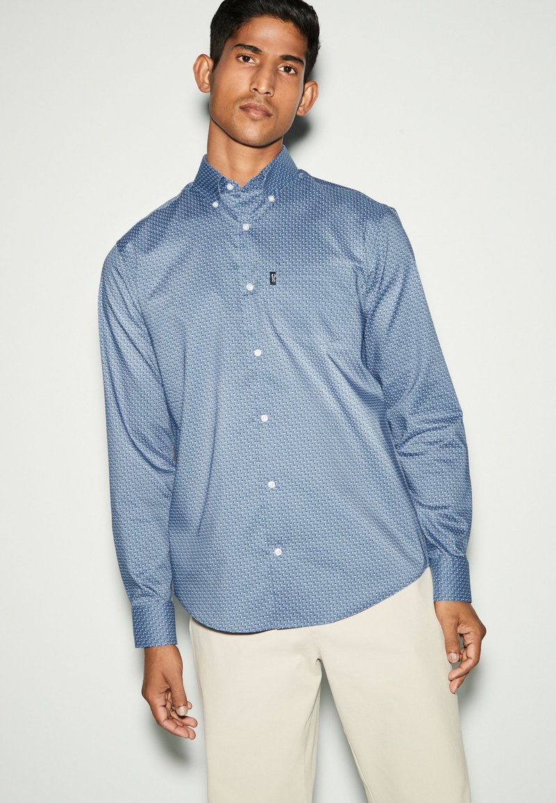 Camicia button-up a maniche lunghe con motivo blu, caratterizzata da un piccolo logo sul petto e un orlo arrotondato, abbinata a dei pantaloni chiari.