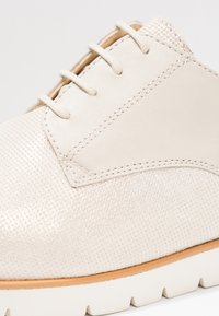 Chaussure beige clair en matériau texturé, avec un bout de chaussure lisse et une semelle blanche contrastante. Coutures détaillées et design à lacets.