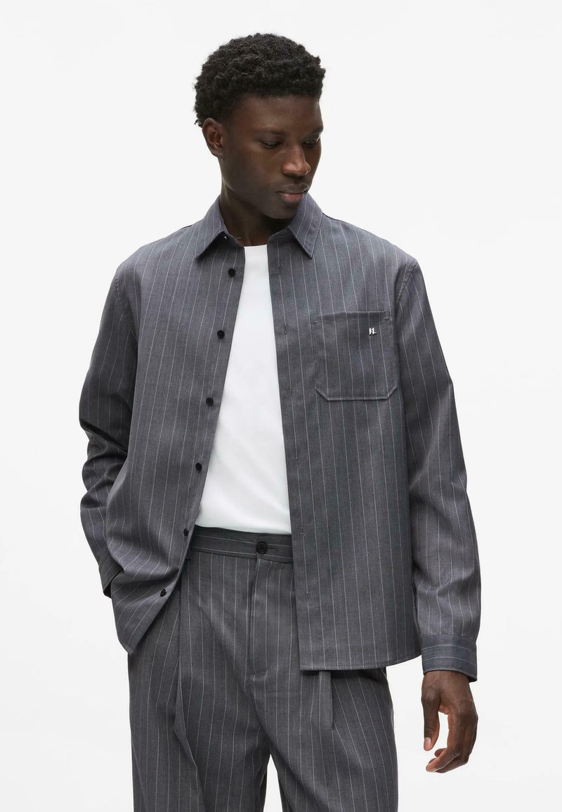 Homme portant une chemise grise à fines rayures boutonnée et un pantalon assorti par-dessus un T-shirt blanc, regardant vers le bas et sur le côté.
