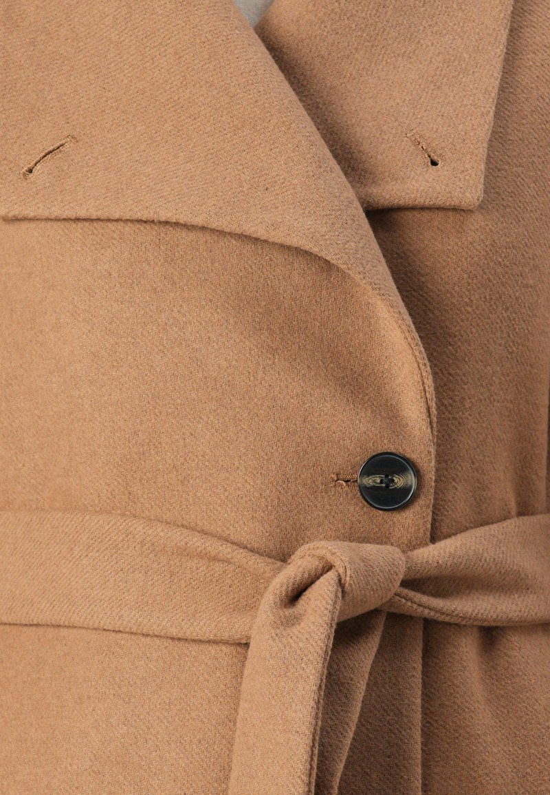 Cappotto in misto lana color sabbia con colletto a risvolto, chiusura a bottoni e cintura annodata in vita. Tessuto morbido e colore neutro e caldo.