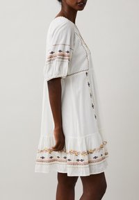 Vitt bomullsklänning med korta puffärmar, prydd med färgglad broderi av blommor och volangkant. Rakt snitt med dekorativ sömnad.