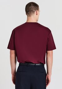 Uomo di spalle che indossa una camicia semplice a maniche corte color bordeaux infilata in pantaloni neri con una cintura nera, su uno sfondo bianco.