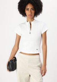 Maglietta bianca a coste con collo zip e maniche corte, caratterizzata da una vestibilità cropped e un piccolo logo nero all'orlo. Abbinata a pantaloni beige e una borsa nera.