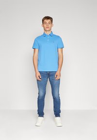 Polo bleu clair avec col et manches courtes, associé à un jean slim bleu et des baskets blanches. Design simple sans motifs.
