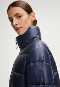 Giacca puffer blu navale con colletto alto, finitura lucida e design trapuntato; include una toppa del logo sulla spalla sinistra.