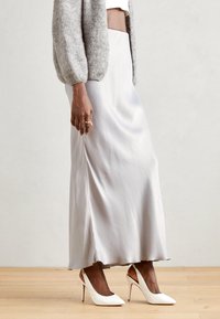 Fustă maxi din mătase, de un gri deschis, cu o textură fină și o deschidere laterală, asortată cu pantofi cu toc înalt și curea albă. Design simplu și elegant.