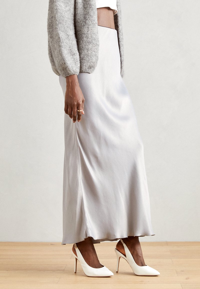 Fustă maxi din mătase, de un gri deschis, cu o textură fină și o deschidere laterală, asortată cu pantofi cu toc înalt și curea albă. Design simplu și elegant.