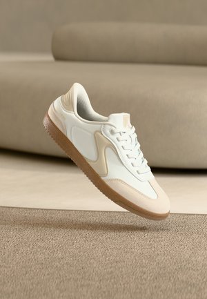 Balts un bēšs ikdienas sneakeris ar gumijas zoli, peldošs virs teksturēta brūna paklāja minimālistiskā bēšā telpā.