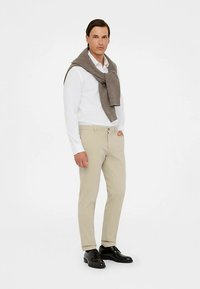 Pantaloni croiți de culoare deschisă, cămașă albă cu nasturi și un pulover gri tricotat așezat peste umeri. Pantofi eleganți negri completează ținuta.