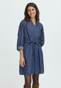 Leende kvinna som bär en knälång denimklänning med uppvikta ärmar, framknappar, knyts midjebälte, svarta strumpbyxor och bruna stövlar.